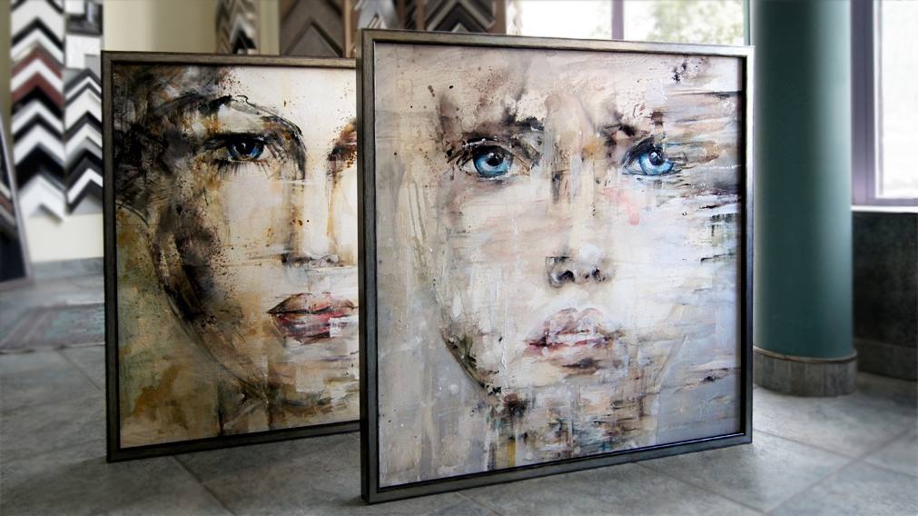 Dwa obrazy w metalowych ramach przedstawiające abstrakcyjne portrety kobiet z intensywnie niebieskimi oczami, ustawione na podłodze z szarych płytek, z widocznymi ramami obrazów w tle i zielonym...