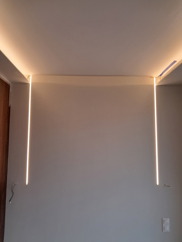 Minimalistyczne oświetlenie LED wzdłuż krawędzi sufitu i ścian, tworzące prostokątną ramę świetlną w szarym pomieszczeniu, widoczne przewody elektryczne przy jednej ze ścian.