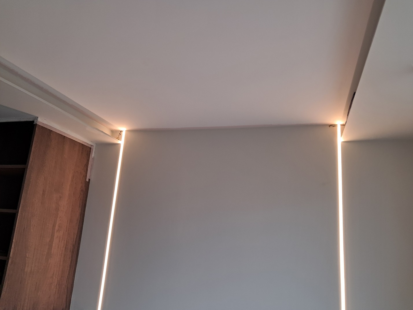 Ukryte oświetlenie LED wzdłuż krawędzi sufitu i ściany, tworzące pionowe linie światła, obok drewnianej szafy.
