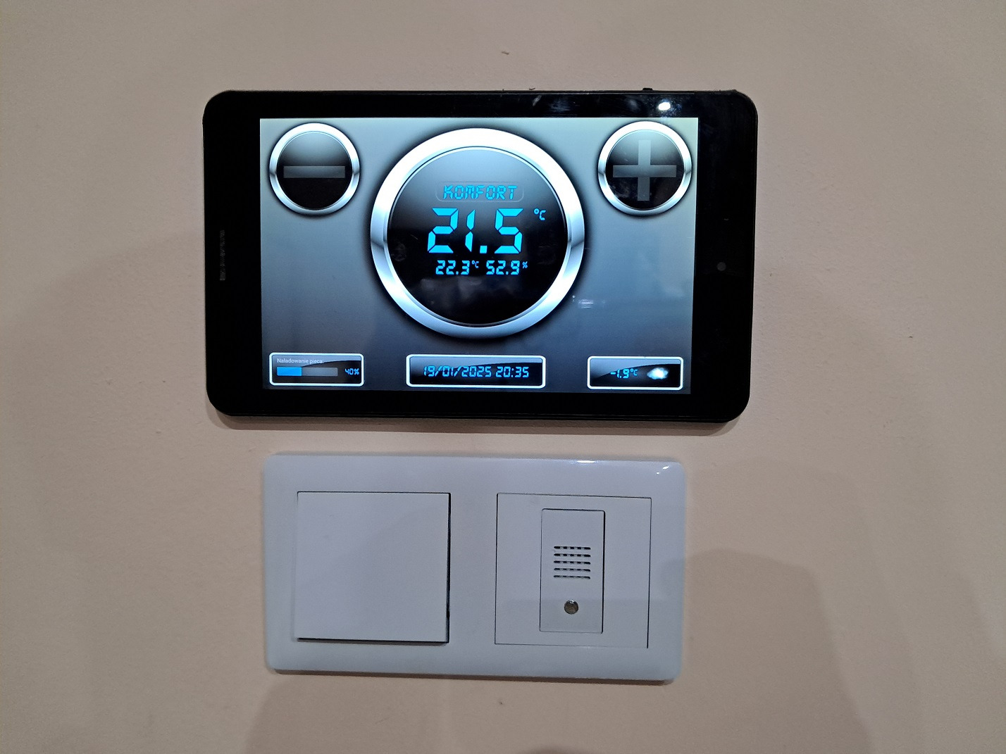 Tablet zamontowany na ścianie, wyświetlający interfejs sterowania temperaturą w pomieszczeniu, obok widoczny biały włącznik światła i panel domofonu.