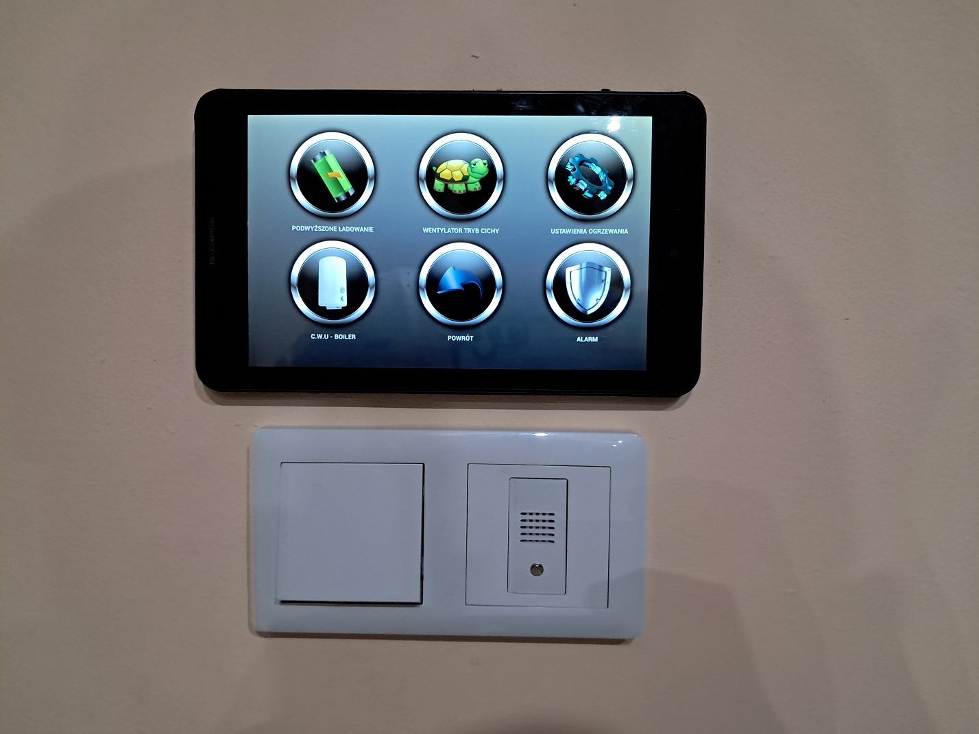 Tablet z interfejsem sterowania systemem domu inteligentnego, zamontowany nad włącznikiem światła i panelem domofonu.