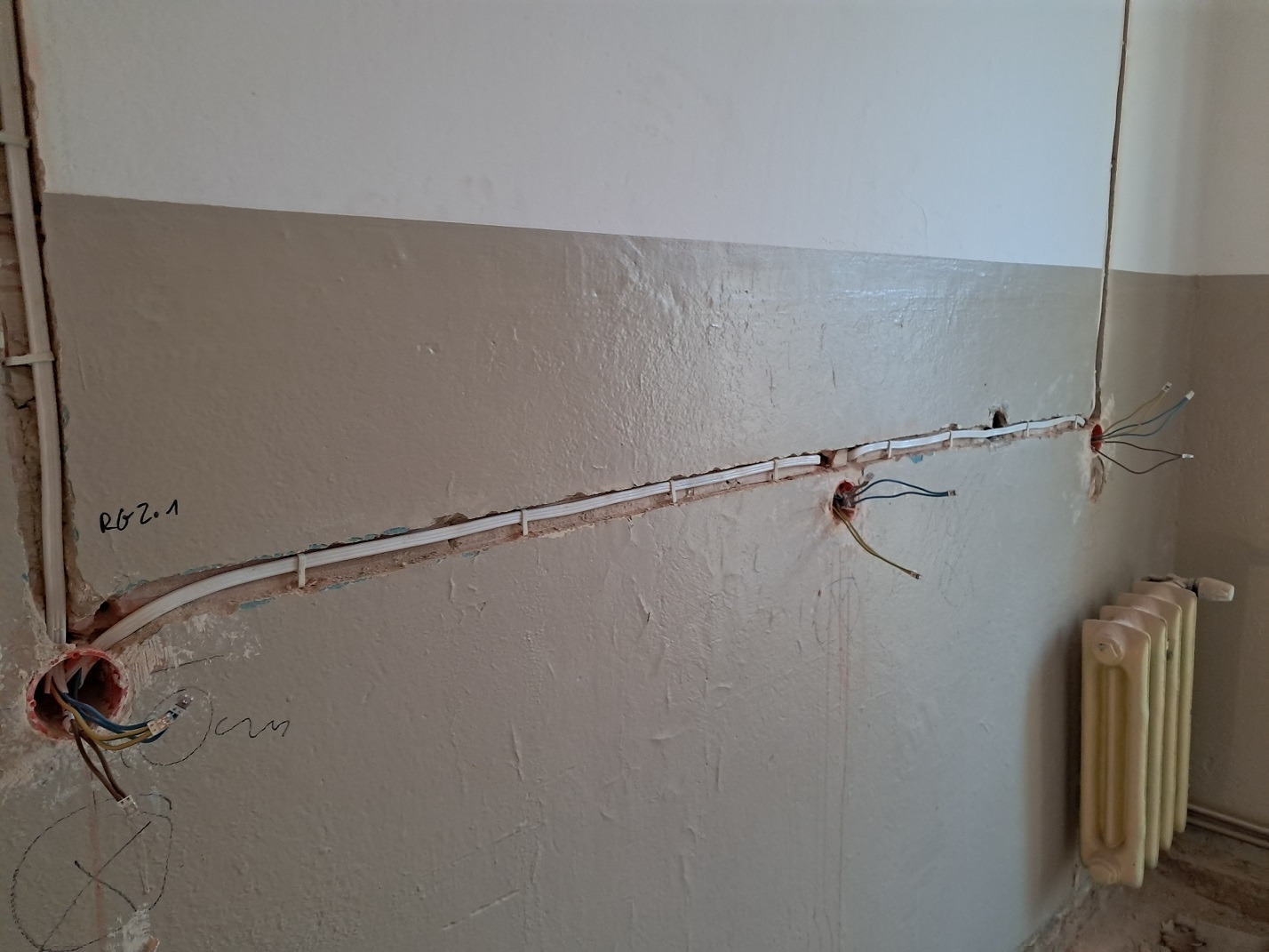 Fragment ściany z bruzdami i puszkami elektrycznymi, w których widoczne są przewody, oraz zamocowanym kablem w białej listwie, obok grzejnik żeberkowy.
