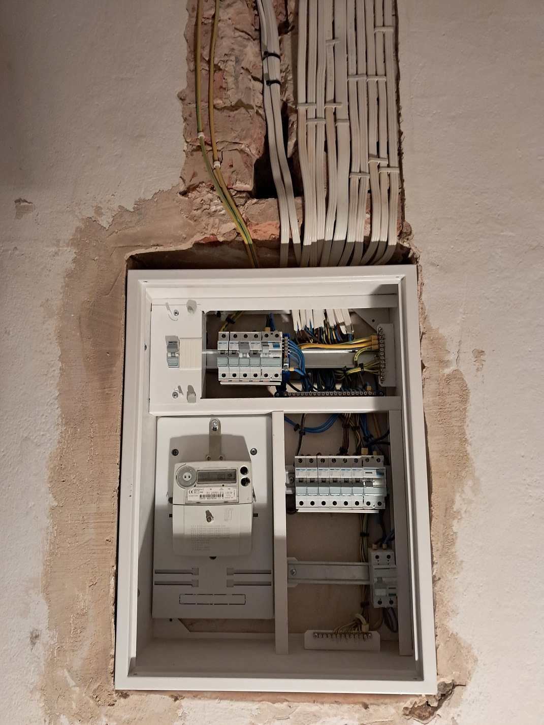 Otwarta skrzynka elektryczna z licznikiem i bezpiecznikami, widoczne kable w ścianie, surowy stan wykończenia ściany.