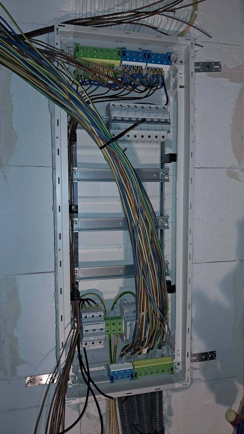 Otwarta skrzynka rozdzielcza z dużą ilością przewodów elektrycznych w różnych kolorach, zamocowana na surowej ścianie z betonu komórkowego.