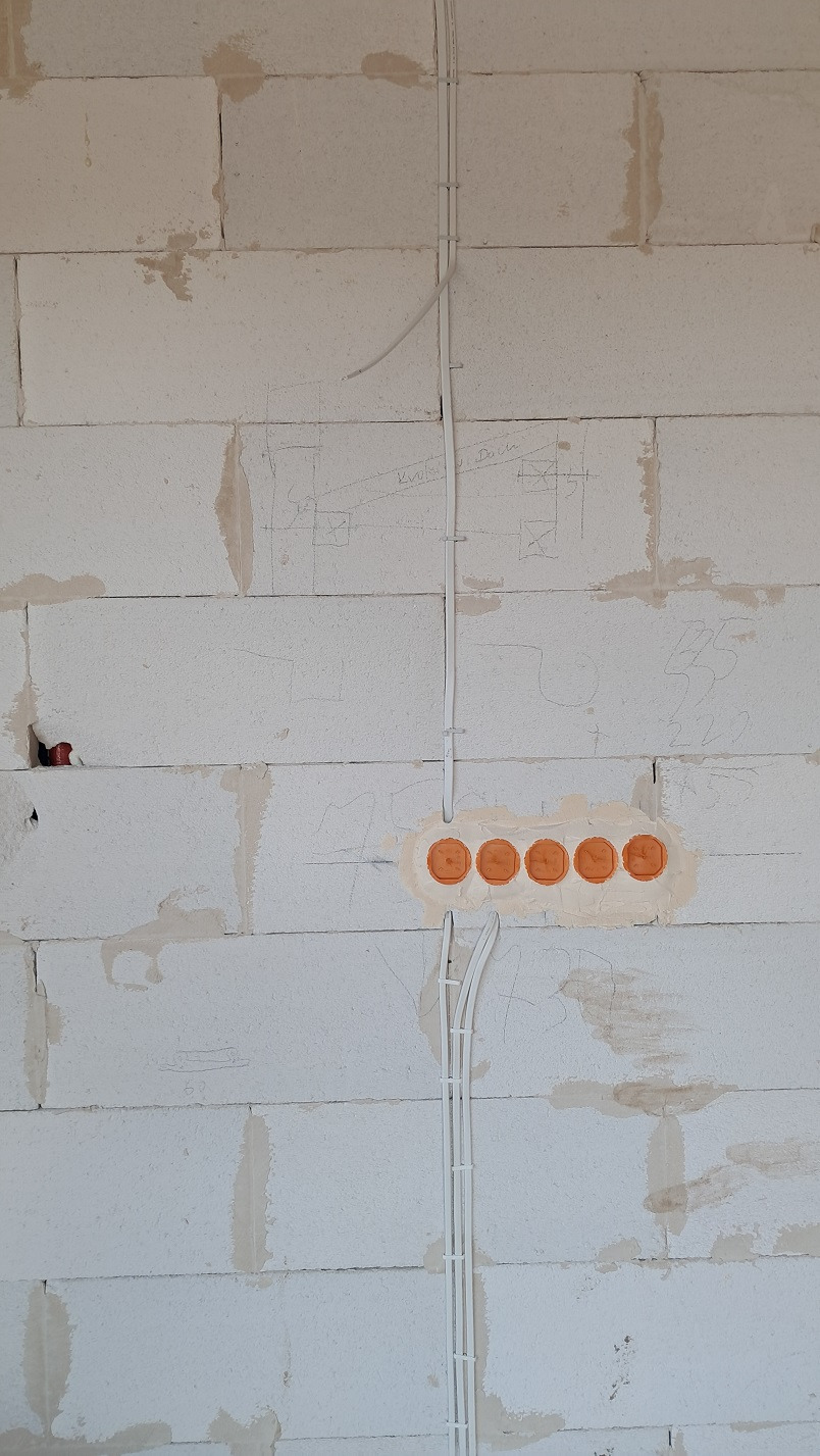 Surowa ściana z betonu komórkowego z widocznymi przewodami elektrycznymi w białych peszlach i pomarańczowymi puszkami pod gniazdka, na ścianie widoczne szkice instalacji.