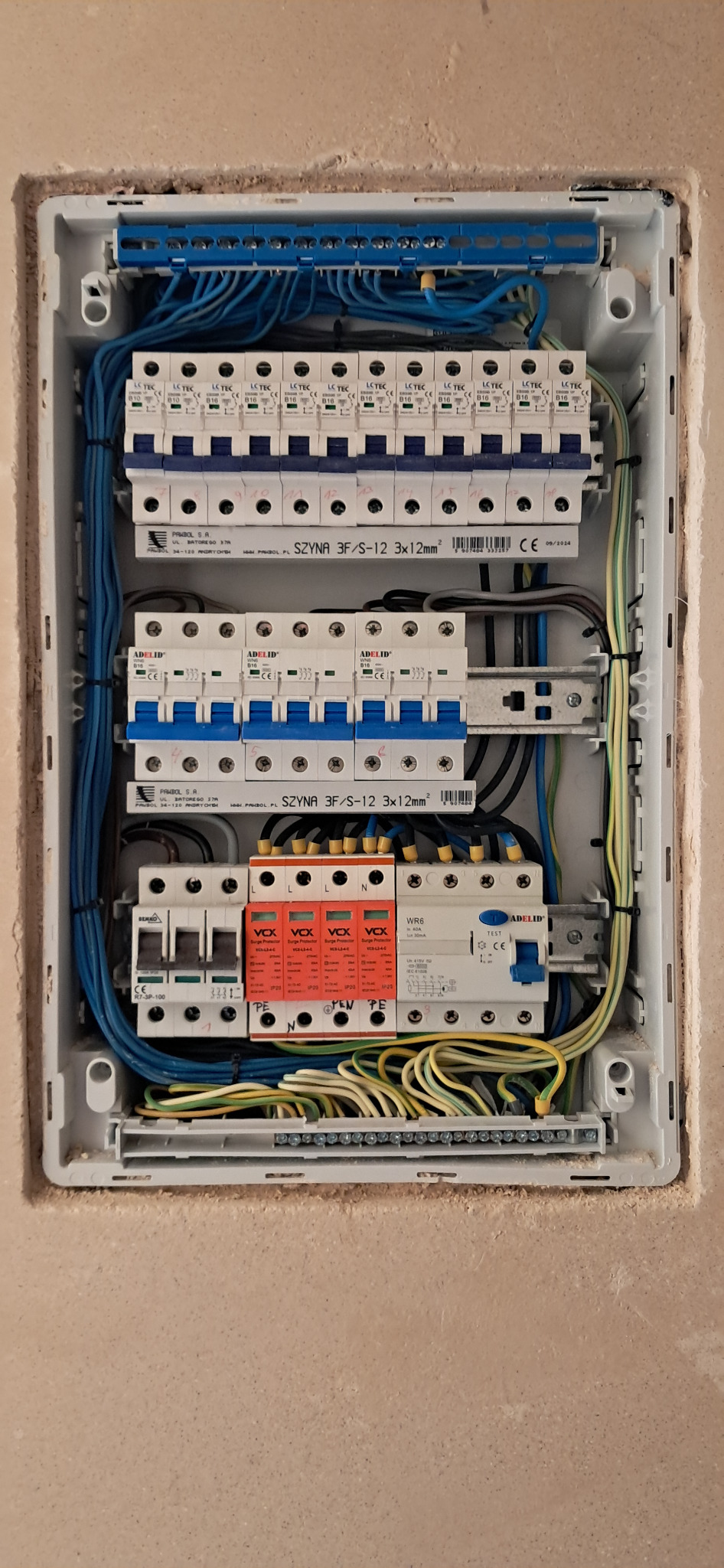 Rozdzielnica elektryczna w obudowie, widoczne zabezpieczenia, przewody i listwy zaciskowe. Instalacja w trakcie montażu, ściana w tle surowa.