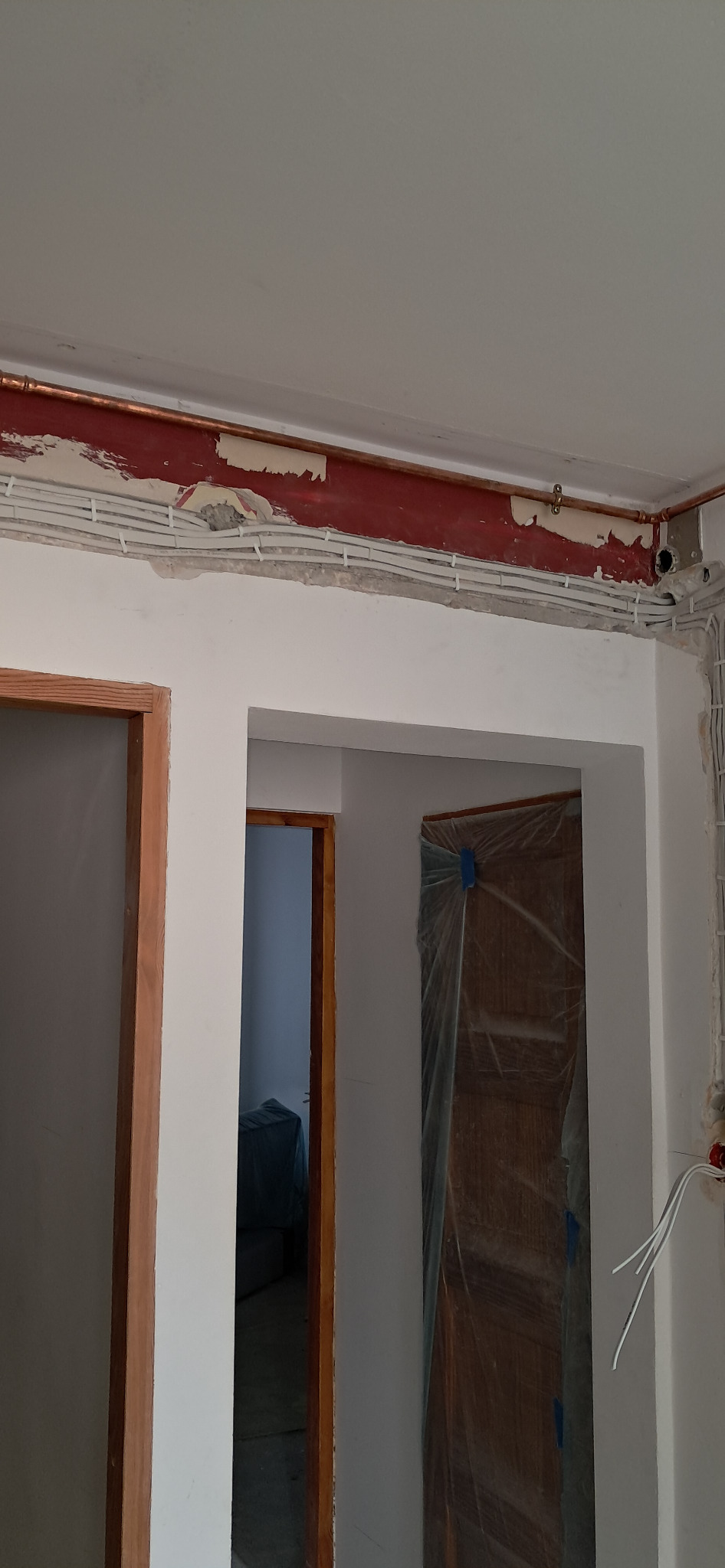 Białe kable elektryczne ułożone wzdłuż ściany pod sufitem, widoczne miedziane rurki i fragmenty czerwonej farby. Drzwi zabezpieczone folią malarską.