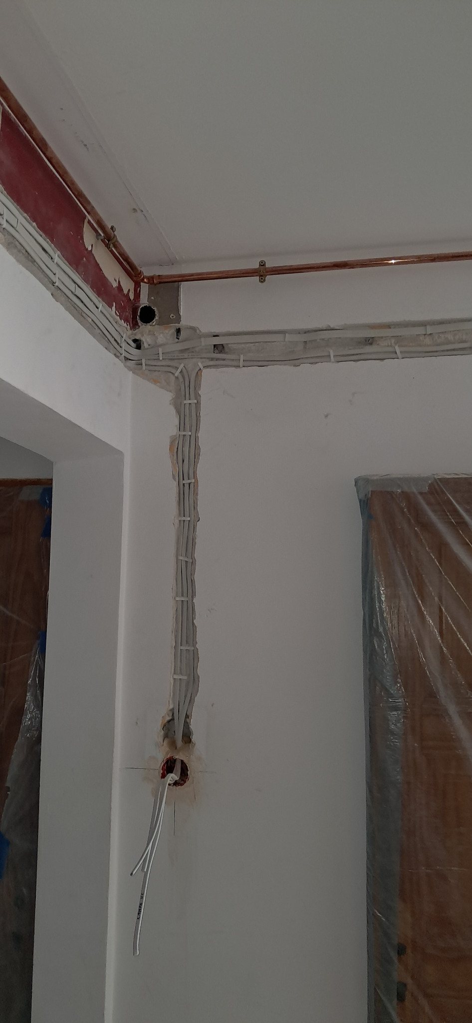 Białe kable elektryczne ułożone w bruzdach na ścianie, zabezpieczone uchwytami. Widoczne rury miedziane pod sufitem i otwór w ścianie z wystającymi przewodami.