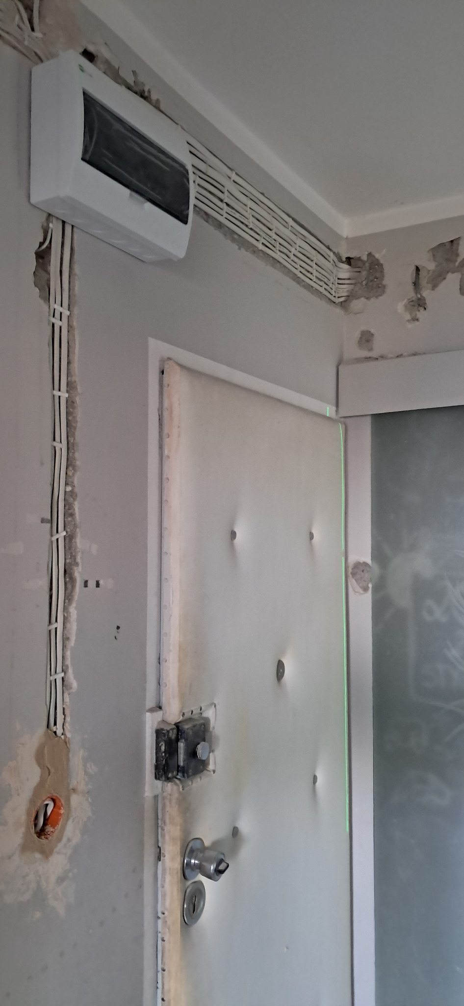 Biała skrzynka elektryczna i kable w korytkach na ścianie w trakcie remontu. Widoczne ślady po kuciu i instalacji. Drzwi obok z metalowymi okuciami.