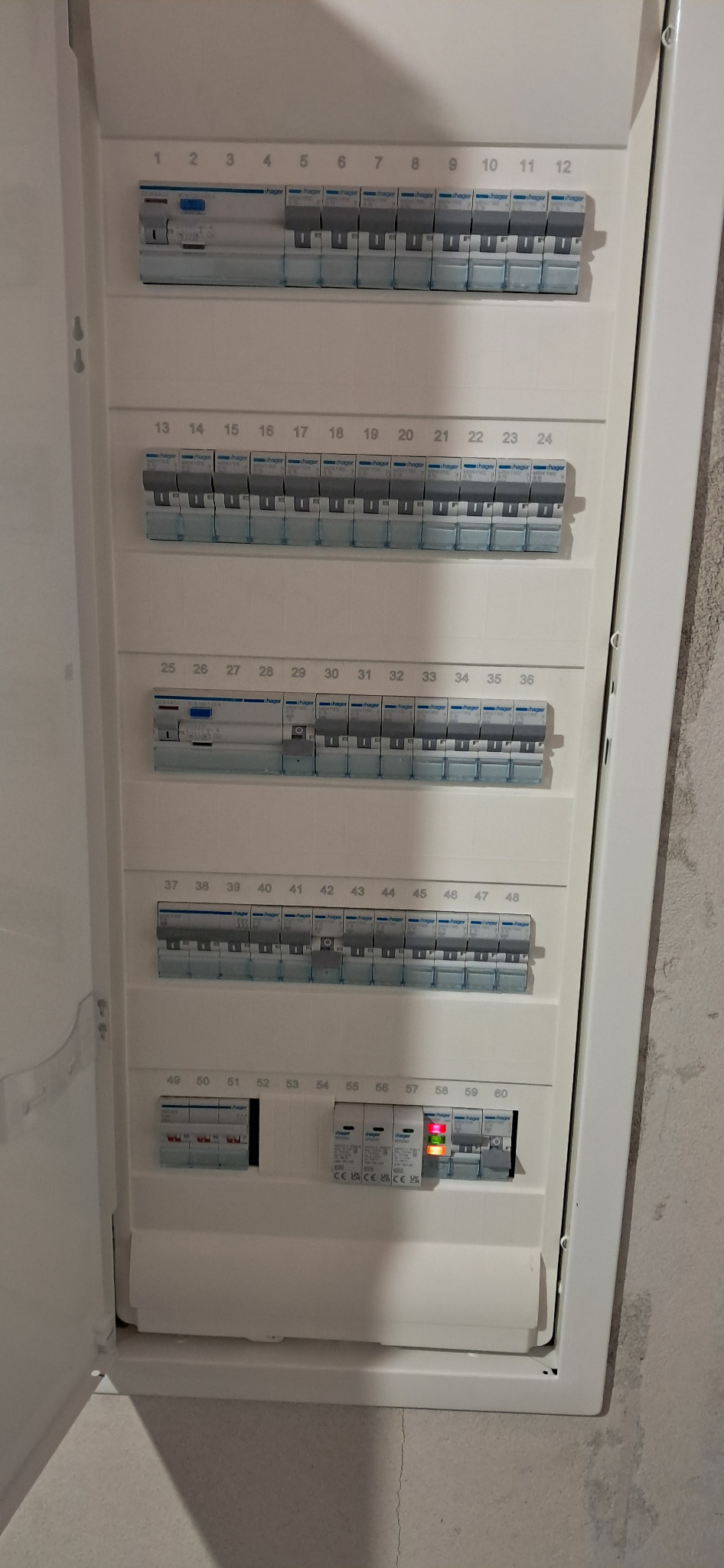 Otwarta skrzynka bezpiecznikowa Hager z numerowanymi wyłącznikami, niektóre w pozycji włączonej, widoczne oznaczenia CE. Biała obudowa na tle surowej ściany.