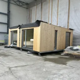 Gotowy modułowy dom z jasnego drewna z dużymi przesuwnymi oknami, umieszczony na kółkach w hali produkcyjnej.