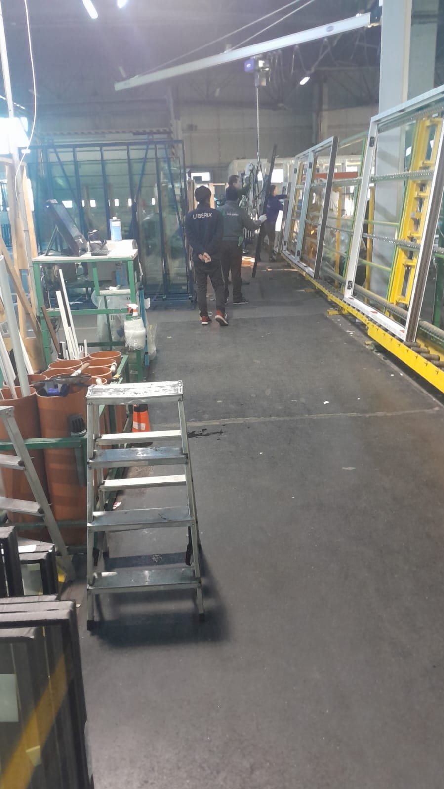Wnętrze hali produkcyjnej z pracownikami w uniformach firmy LIBERIX, produkcja okien, drabina na pierwszym planie, okna ustawione w rzędzie w tle.