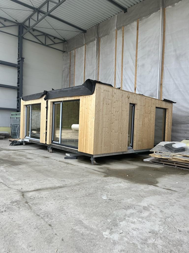 Gotowy modułowy dom z jasnego drewna z dużymi przesuwnymi oknami, umieszczony na kółkach w hali produkcyjnej.