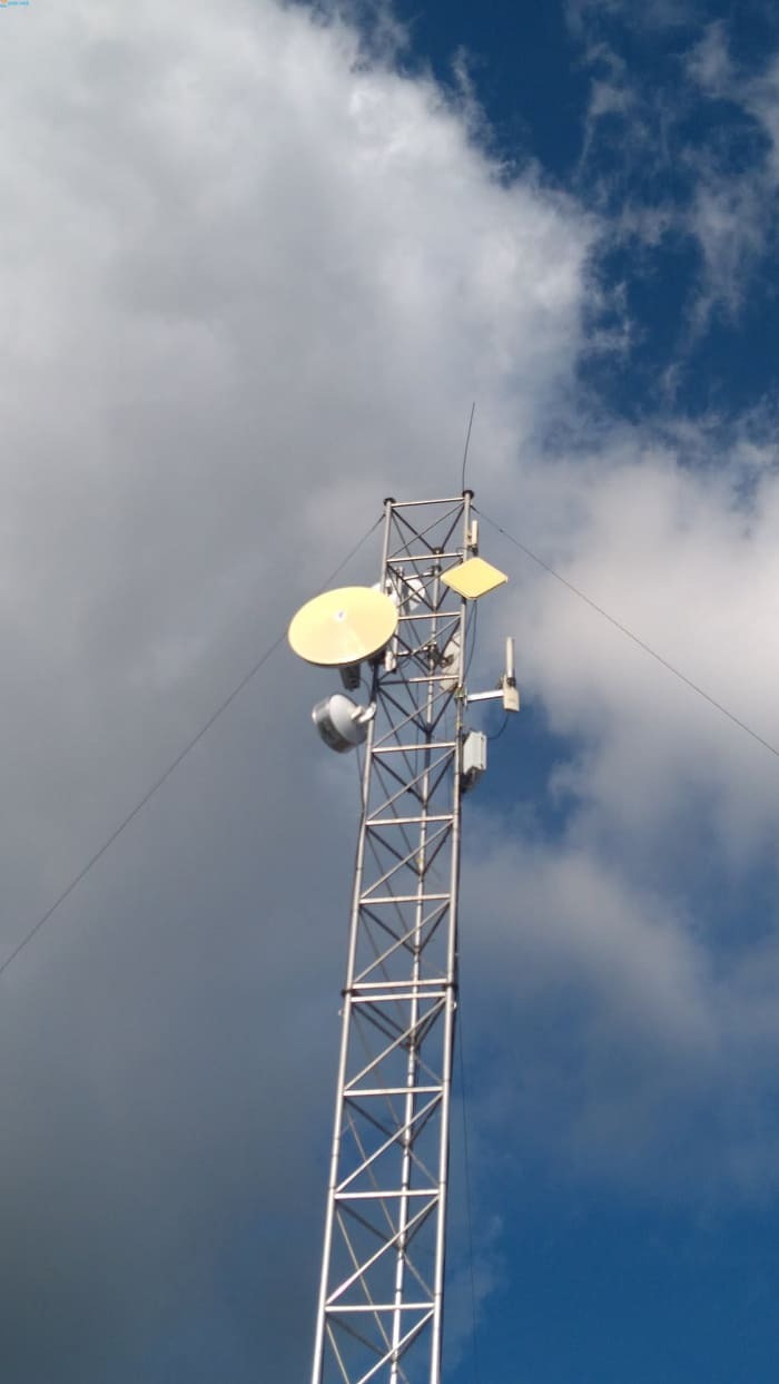 Metalowa wieża telekomunikacyjna z antenami parabolicznymi i panelowymi na tle błękitnego nieba z chmurami.