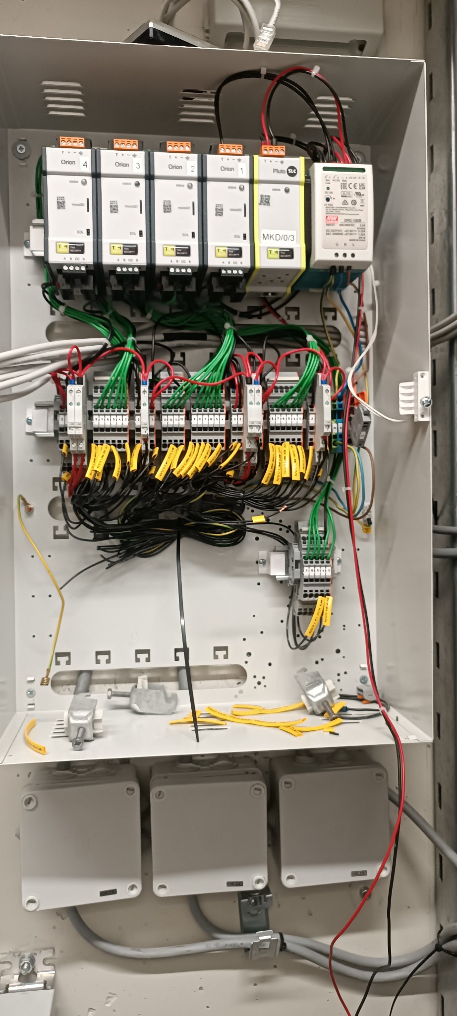 Otwarta szafa sterownicza z modułami Orion 1-4 i Pluto SLC, licznymi przewodami w kolorach zielonym, żółtym i czarnym, oraz szynami DIN, widoczne również trzy szare puszki elektryczne na dole.