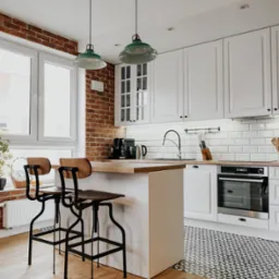 Jasna kuchnia z wyspą, białymi szafkami, ceglaną ścianą i drewnianymi podłogami, oświetlona dwoma zielonymi lampami wiszącymi nad wyspą.