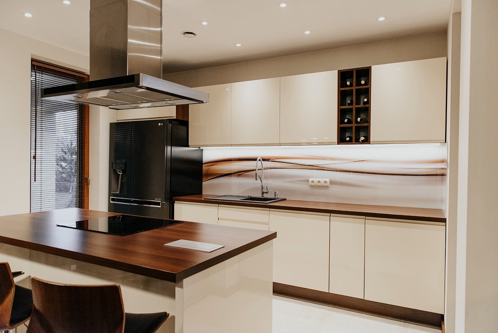 Nowoczesna kuchnia z beżowymi, lakierowanymi frontami, drewnianym blatem i wyspą z płytą indukcyjną oraz czarną lodówką side-by-side i wbudowaną półką na wino.