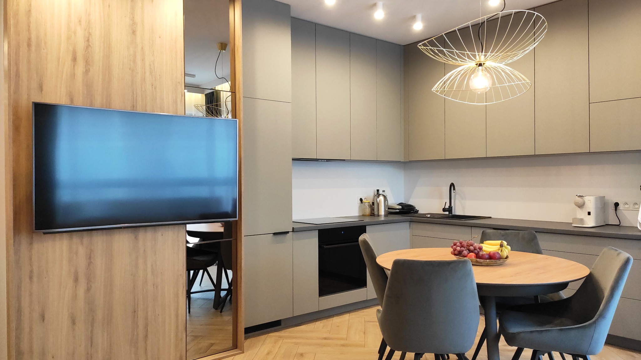 Nowoczesna kuchnia w odcieniach szarości z drewnianymi akcentami, okrągłym stołem i dekoracyjną lampą wiszącą. Na blacie widoczny owocowy poczęstunek.