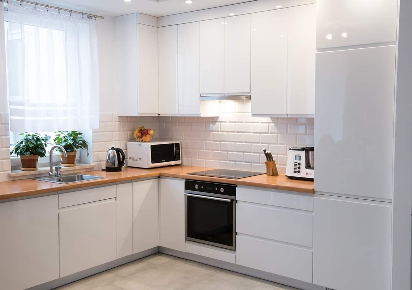 Jasna, nowoczesna kuchnia z białymi, lakierowanymi frontami, drewnianym blatem i białymi płytkami na ścianie. Na blacie widoczny czajnik, mikrofalówka i toster.