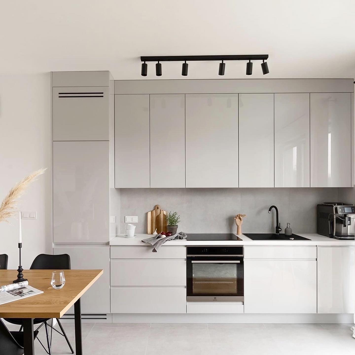 Nowoczesna, minimalistyczna kuchnia w odcieniach szarości z błyszczącymi frontami, oświetleniem punktowym i drewnianym stołem z czarnymi krzesłami w tle.