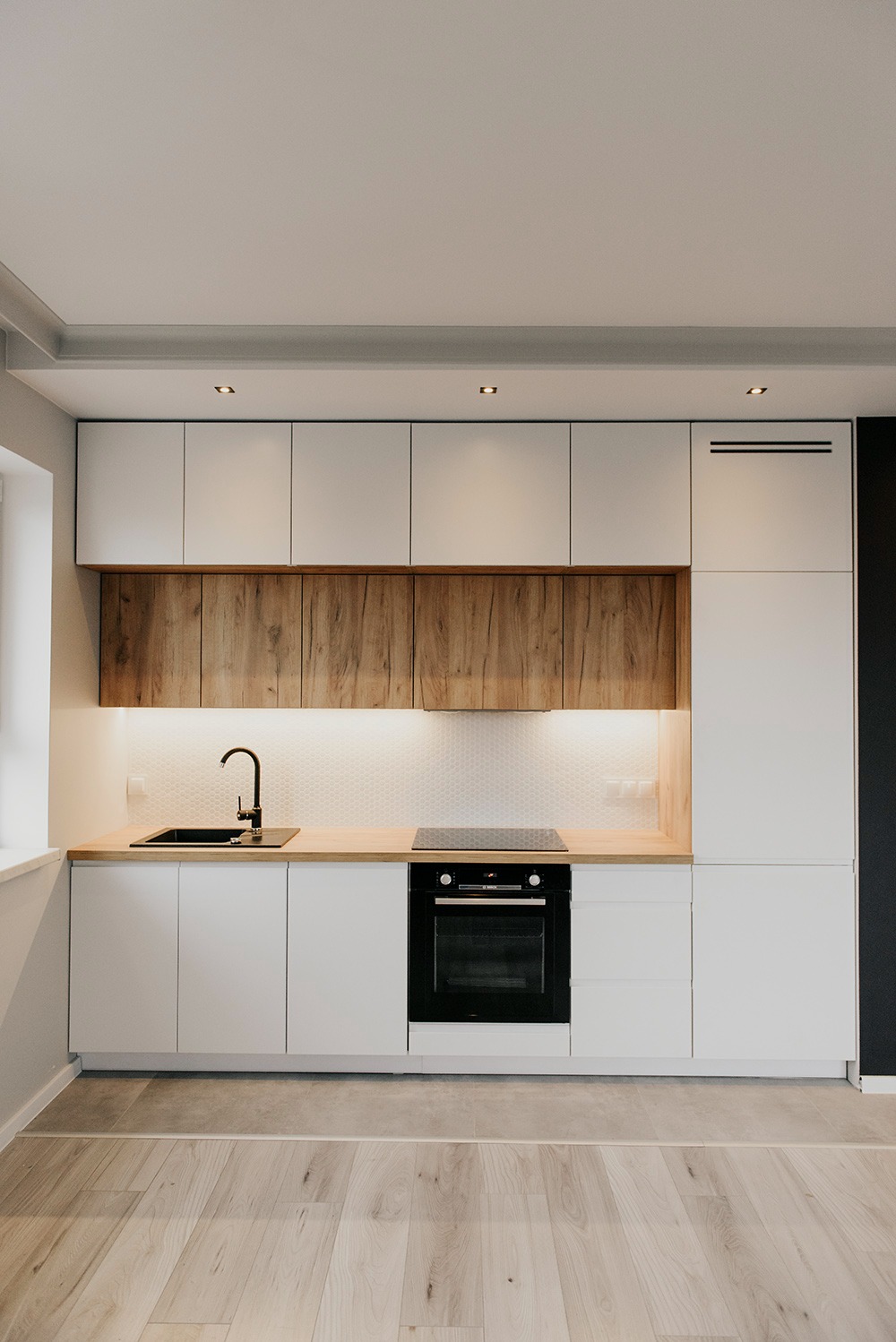 Nowoczesna kuchnia z białymi szafkami i drewnianymi frontami górnymi, czarnym zlewem i płytą indukcyjną, oświetleniem LED pod szafkami i drewnianym blatem.