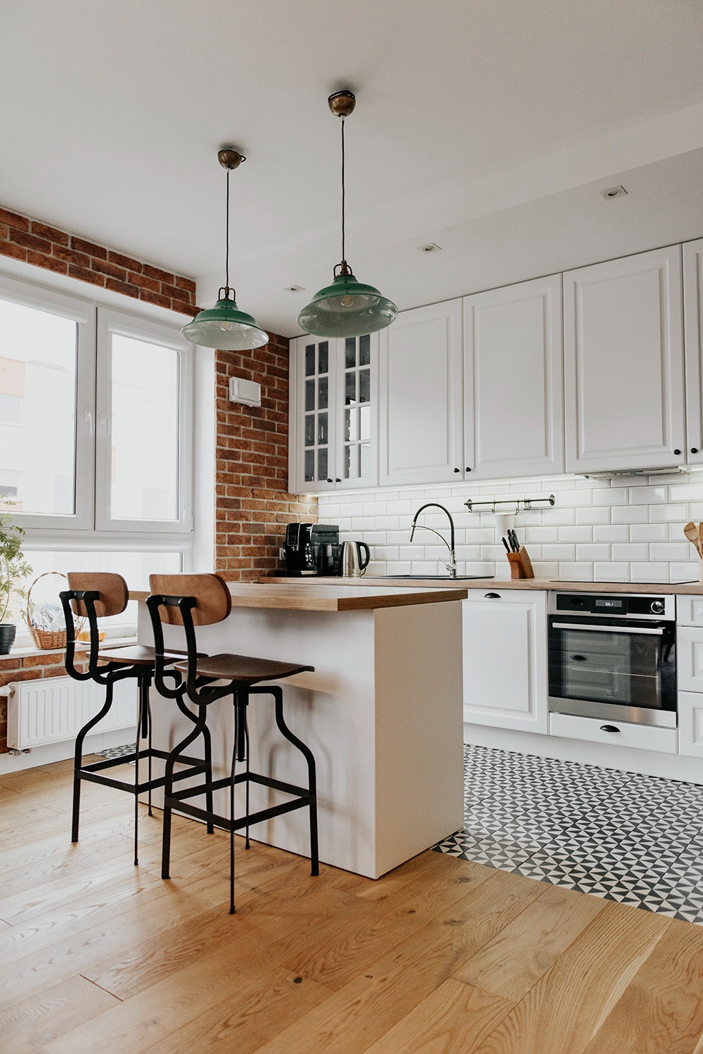 Jasna kuchnia z wyspą, białymi szafkami, ceglaną ścianą i drewnianymi podłogami, oświetlona dwoma zielonymi lampami wiszącymi nad wyspą.