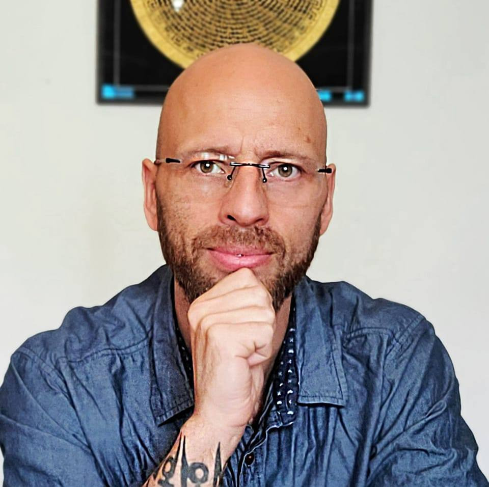 Portret mężczyzny z brodą i okularami, z ręką pod brodą, widoczny tatuaż na przedramieniu, w tle abstrakcyjna grafika.