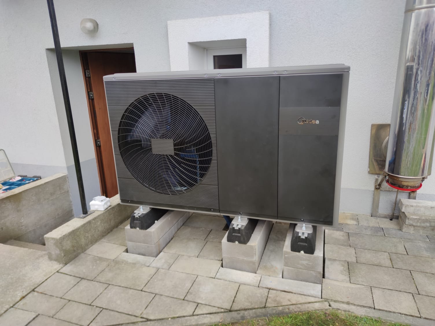 Zewnętrzna jednostka pompy ciepła marki Midea, zamontowana na betonowych blokach przy ścianie budynku z widocznym kominem i schodami.