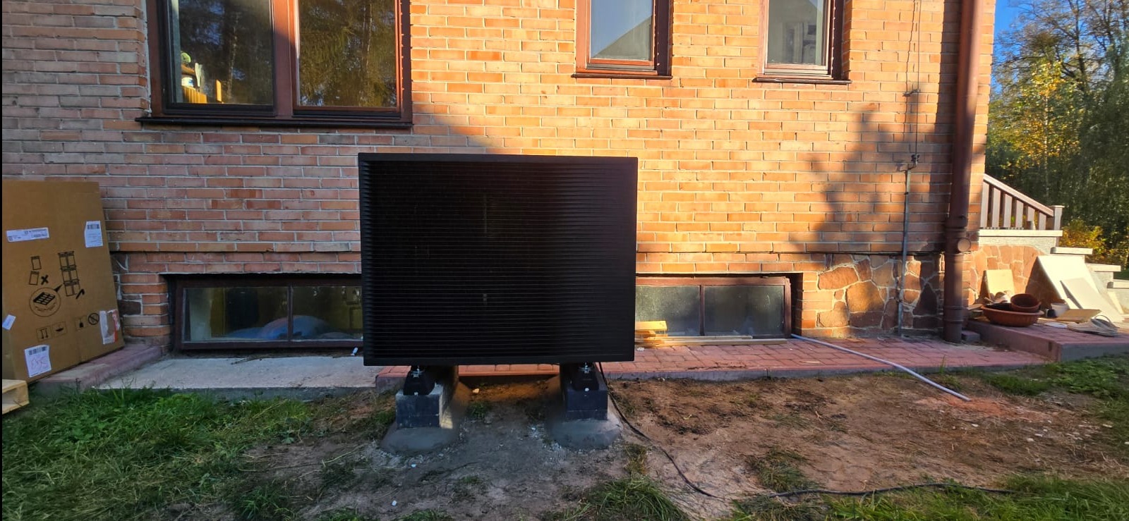 Czarna pompa ciepła powietrze-woda zamontowana na betonowych fundamentach przy ceglanym budynku, widoczne okna piwniczne i kartonowe opakowanie w tle.