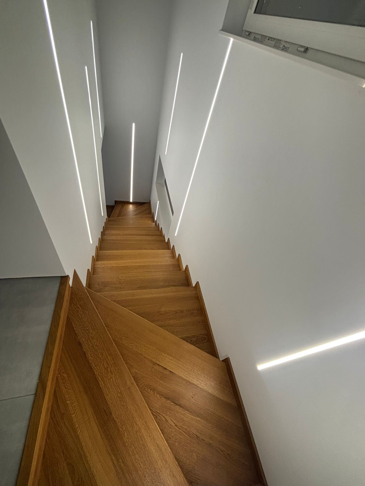 Nowoczesne schody z drewna dębowego, oświetlone pionowymi liniami LED na białych ścianach, widok z góry na dół.
