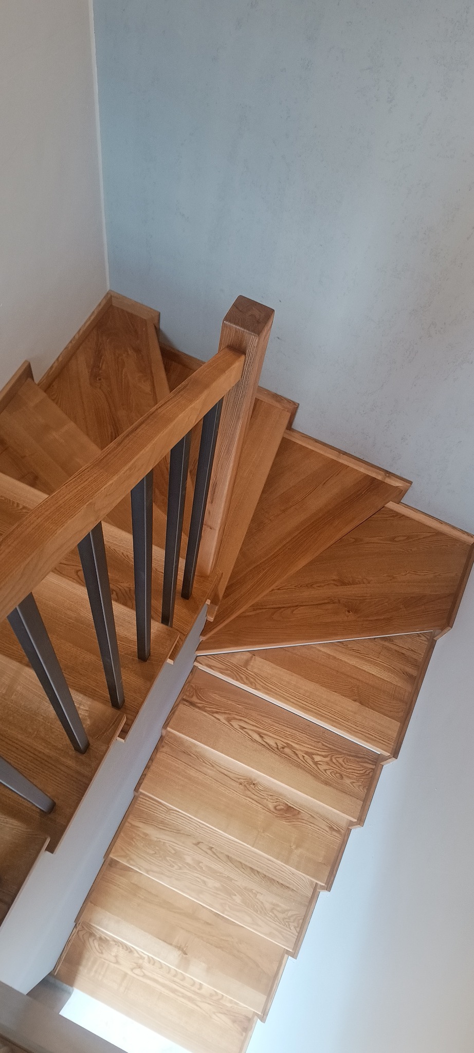 Kątowe ujęcie spiralnych schodów z jasnego drewna z metalowymi balustradami na tle jasnych ścian.