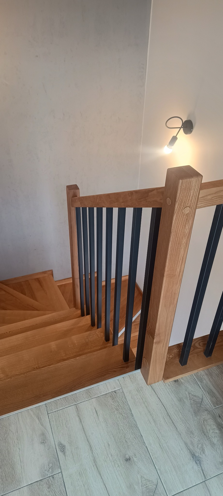 Nowoczesne schody z drewnianymi stopniami i balustradą z drewnianymi słupkami oraz czarnymi, pionowymi elementami, oświetlone lampą ścienną.