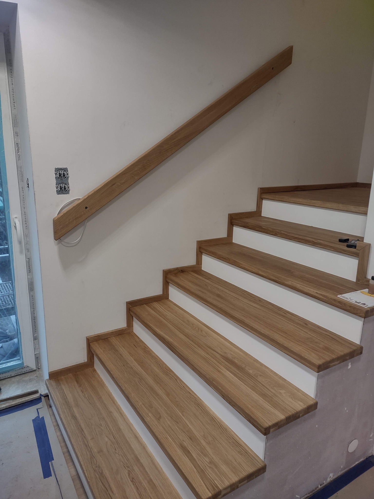 Jasne, dębowe stopnie schodów z białymi podstopnicami i drewnianą poręczą, zamontowaną na ścianie. Widoczne gniazdka elektryczne i fragment okna.