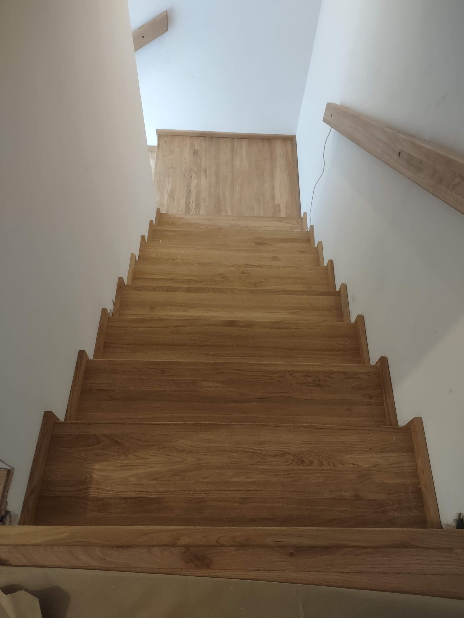 Jasne, drewniane schody z widocznym usłojeniem, prowadzące w górę, z fragmentem drewnianej poręczy po prawej stronie. Minimalistyczne wykończenie ścian.