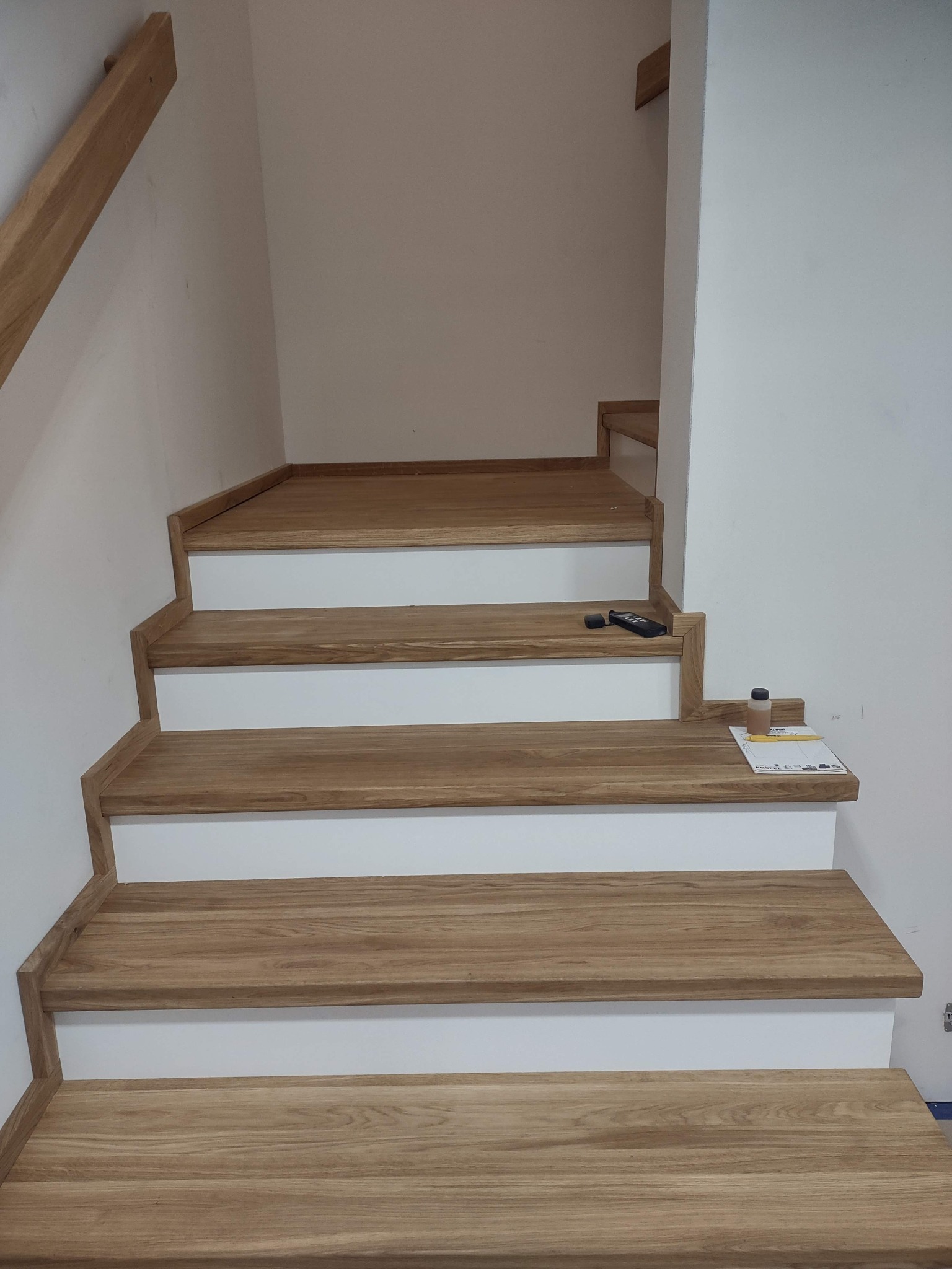 Jasne, dębowe stopnie z białymi podstopnicami w nowoczesnym domu. Na jednym ze stopni leżą dokumenty i pilot, co dodaje autentyczności.