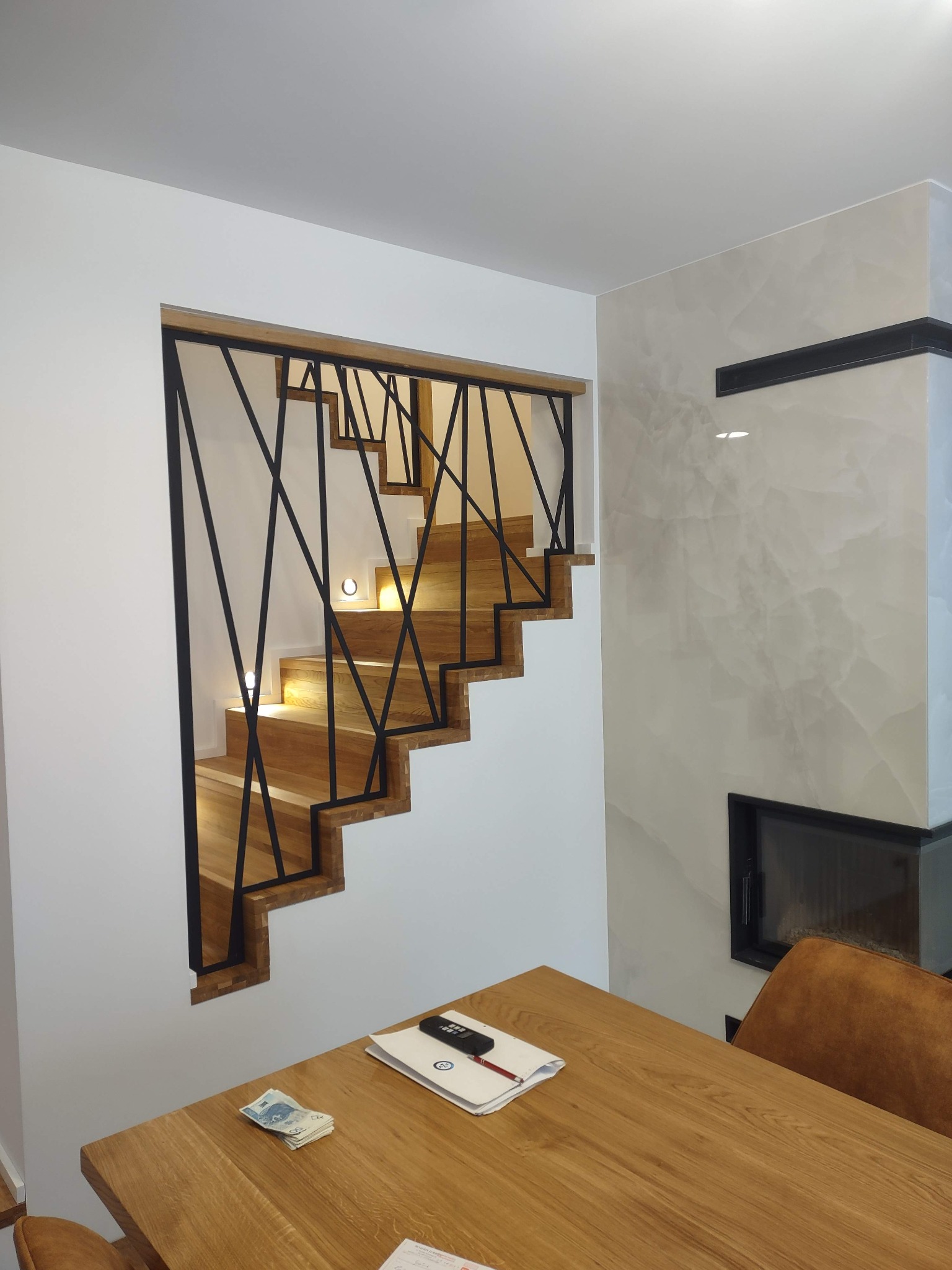 Nowoczesne schody drewniane z czarną, geometryczną balustradą i podświetleniem LED, widoczne zza drewnianego stołu w jasnym wnętrzu.