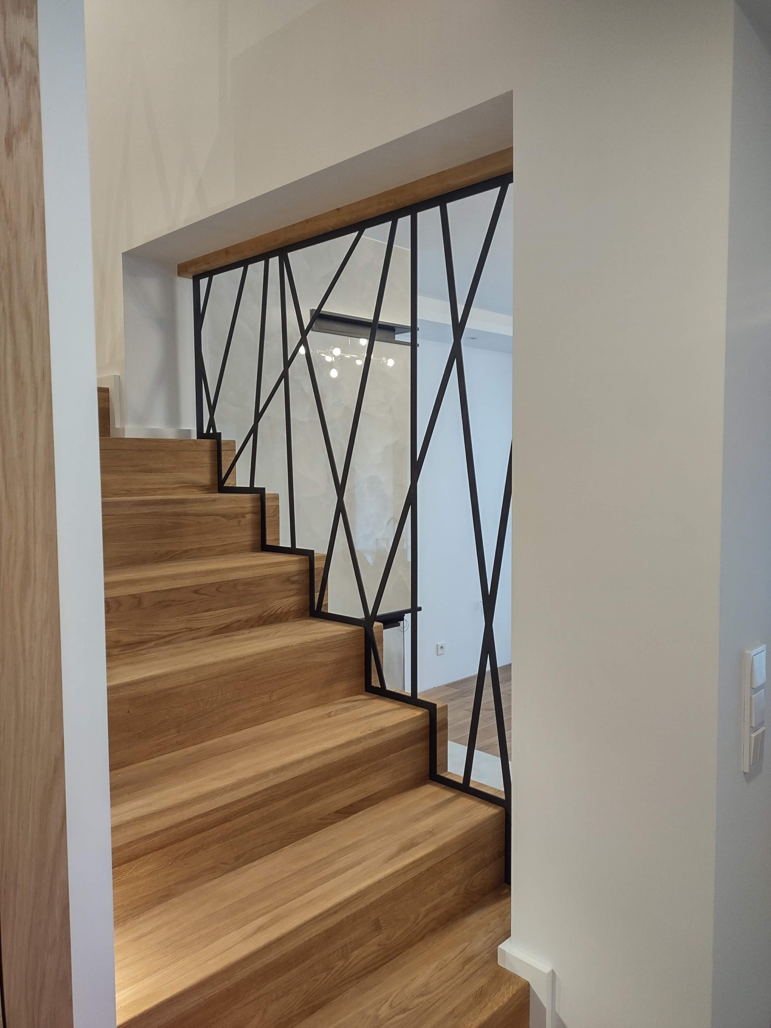 Jasne, drewniane stopnie schodów z czarną, geometryczną balustradą. Nowoczesny design, minimalistyczne wnętrze z białymi ścianami i subtelnym oświetleniem.