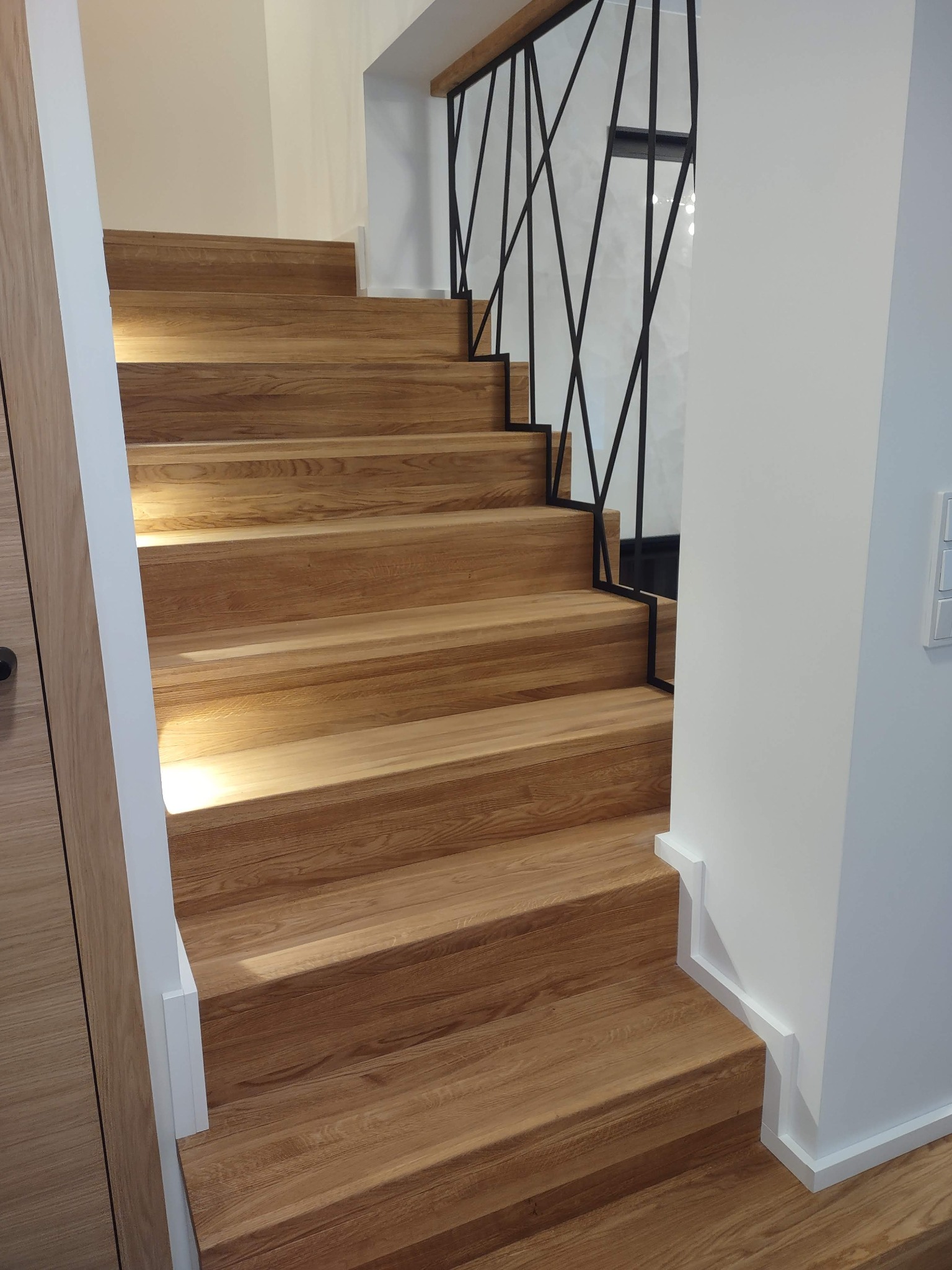 Jasne, dębowe stopnie z podświetleniem LED i czarną, geometryczną balustradą. Nowoczesne schody w białym wnętrzu z drewnianymi akcentami.