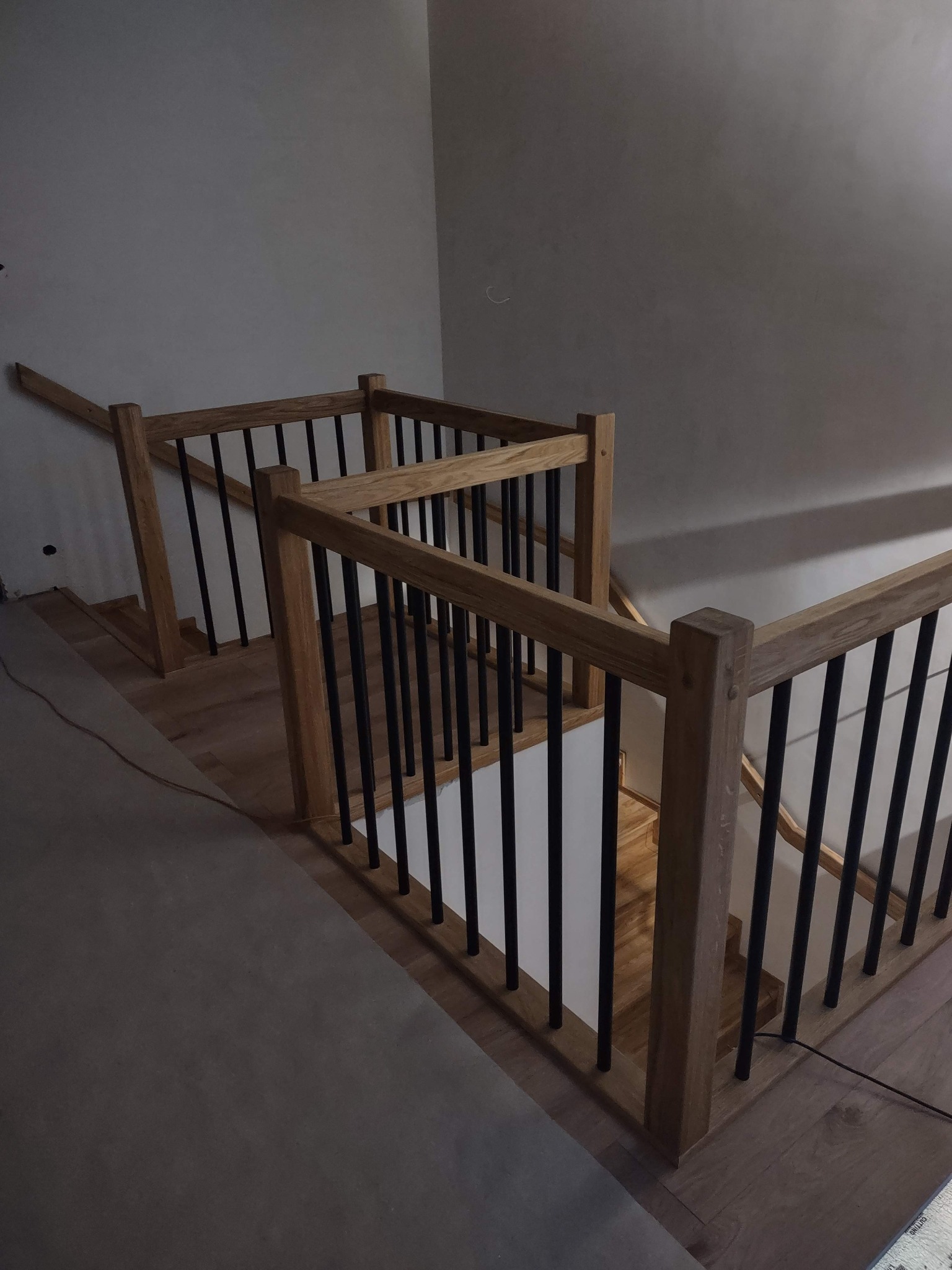 Jasne, dębowe schody z czarnymi, metalowymi balustradami, widziane z góry, w surowym wnętrzu. Widoczne niedokończone ściany i podłogi.