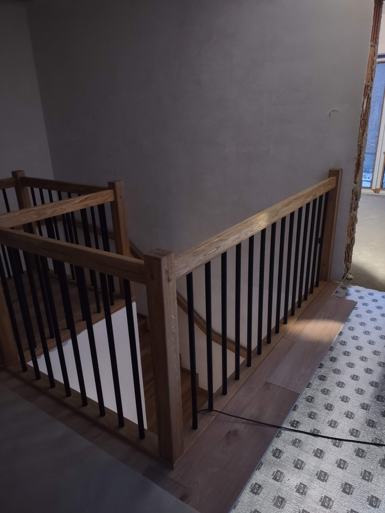 Jasne, drewniane schody z czarnymi balustradami, widok z góry. Częściowo wykończone podłogi i zabezpieczona folią podłoga w tle. Nowoczesny design.