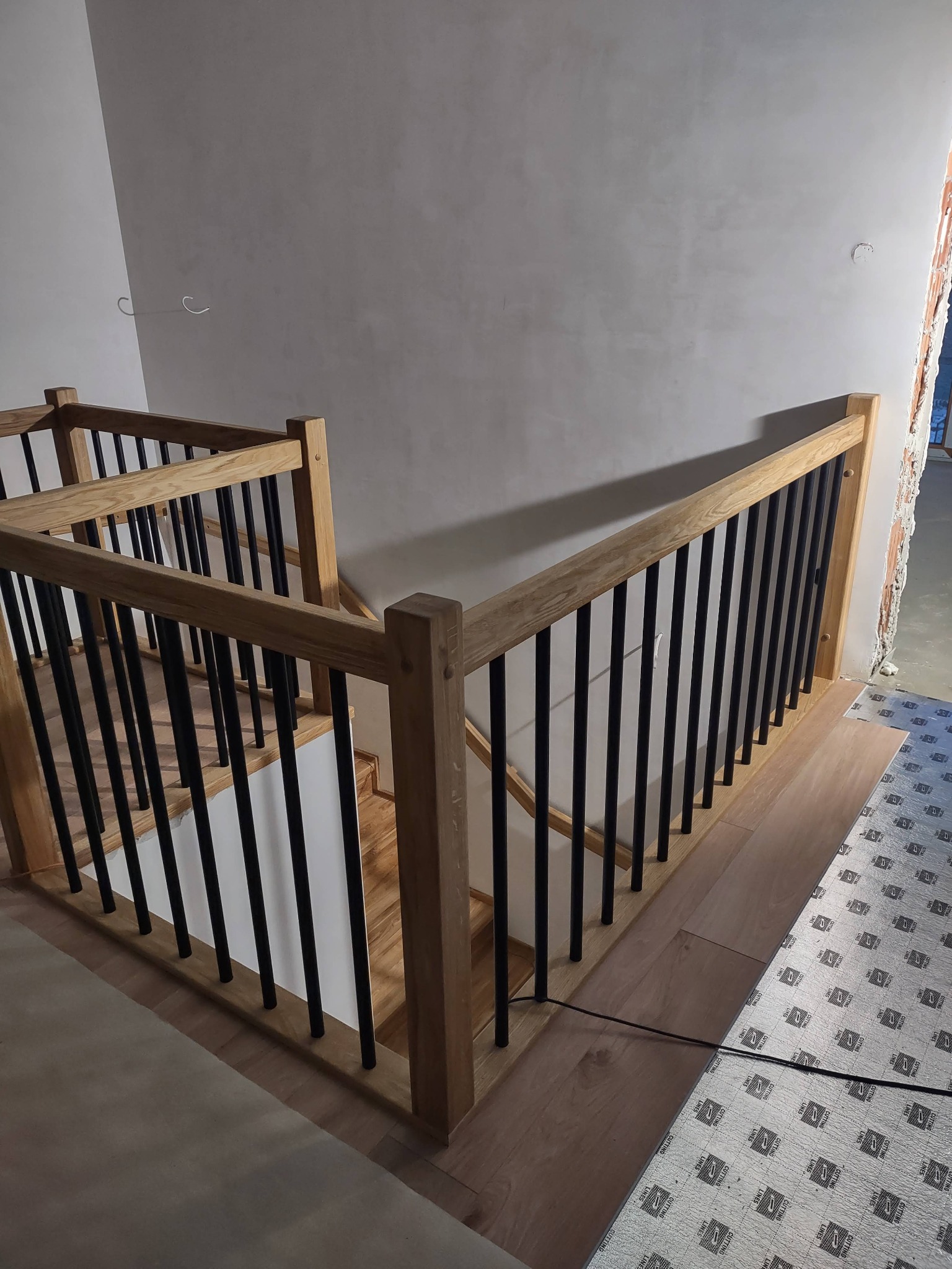 Jasne, dębowe balustrady z czarnymi, pionowymi szczeblami prowadzą w dół. Widoczne fragmenty drewnianych stopni i podłogi. Ściany w surowym stanie.