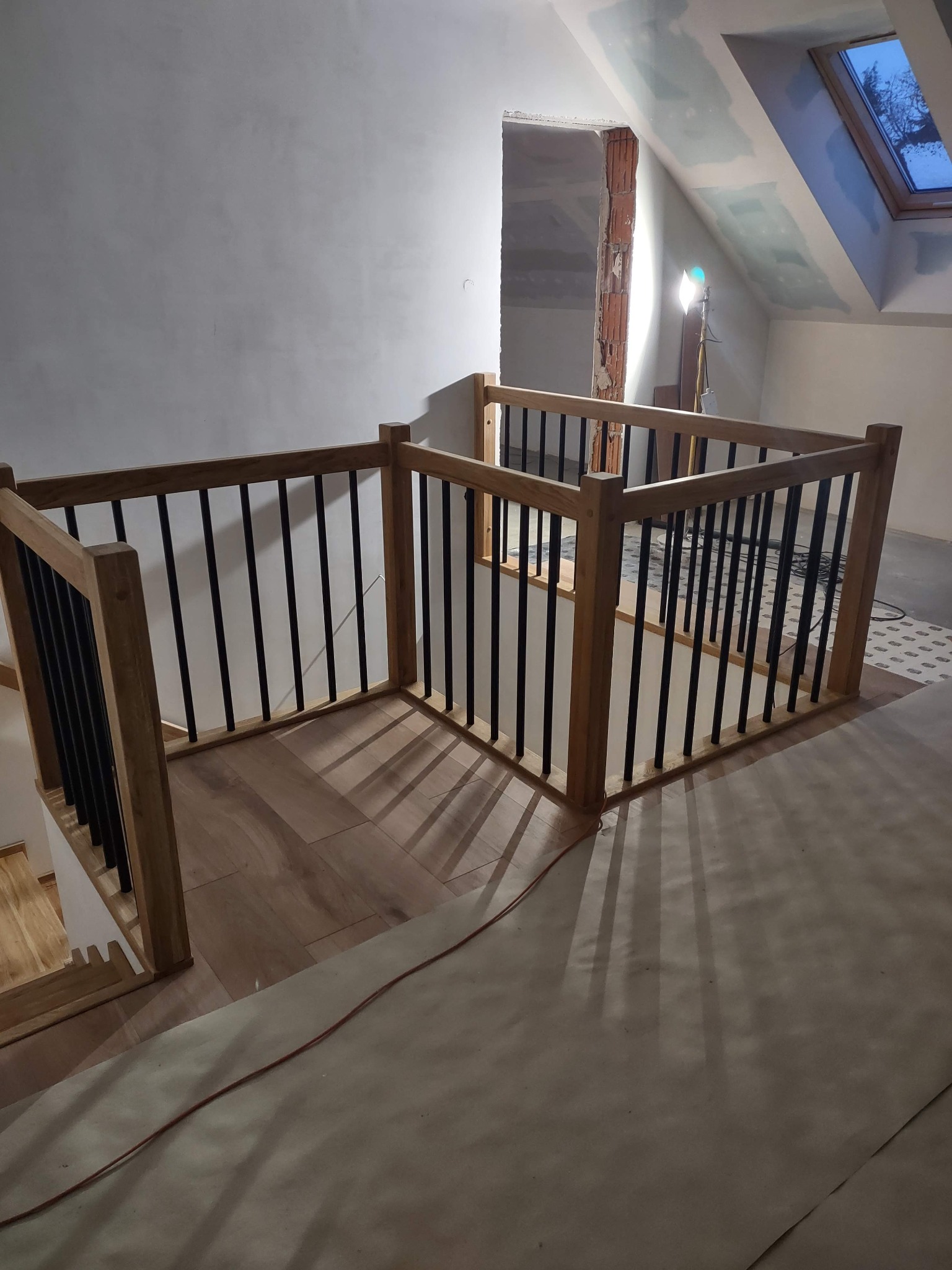 Fragment balustrady z jasnego drewna i czarnymi szczebelkami, zabezpieczającej schody. W tle widoczne okno dachowe i surowy stan wykończenia wnętrza.