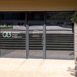 GGK Sp. z o.o. - Wejście do biura rachunkowego z logo Gdańskiej Grupy Księgowych, widoczne godziny otwarcia i dane kontaktowe. Zewnętrzny widok, jasna elewacja i kostka brukowa.
