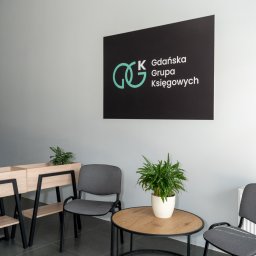 GGK Sp. z o.o. - Wnętrze biura: minimalistyczny design z szarymi krzesłami, drewnianym stolikiem z rośliną i logo 'Gdańska Grupa Księgowych' na ścianie. Stonowana kolorystyka.