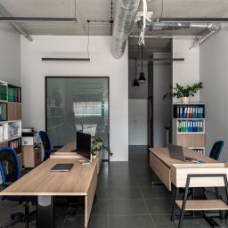 GGK Sp. z o.o. - Nowoczesne biuro z dwoma biurkami, laptopami, regałami z segregatorami i roślinami doniczkowymi. Minimalistyczny design z szarą podłogą i betonowym sufitem.