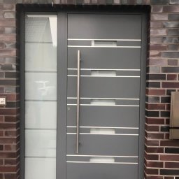 POŻAROWSZCZYK DARIUSZ P.H.U. DARMEX - Nowoczesne szare drzwi wejściowe z aluminiową ramą i pionowym uchwytem, z bocznym panelem ze szkła matowego, osadzone w murze z cegły klinkierowej, z wycieraczką 'Welcome'.