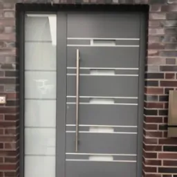 Nowoczesne szare drzwi wejściowe z aluminiową ramą i pionowym uchwytem, z bocznym panelem ze szkła matowego, osadzone w murze z cegły klinkierowej, z wycieraczką 'Welcome'.