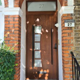 POŻAROWSZCZYK DARIUSZ P.H.U. DARMEX - Nowoczesne, brązowe drzwi wejściowe z przeszkleniami i długim, czarnym uchwytem, numer domu '27' nad drzwiami i obok, kamera monitoringu widoczna nad drzwiami.