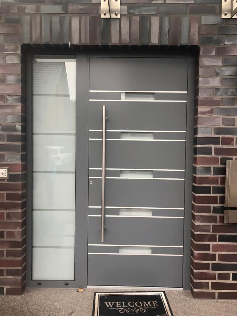 Nowoczesne szare drzwi wejściowe z aluminiową ramą i pionowym uchwytem, z bocznym panelem ze szkła matowego, osadzone w murze z cegły klinkierowej, z wycieraczką 'Welcome'.