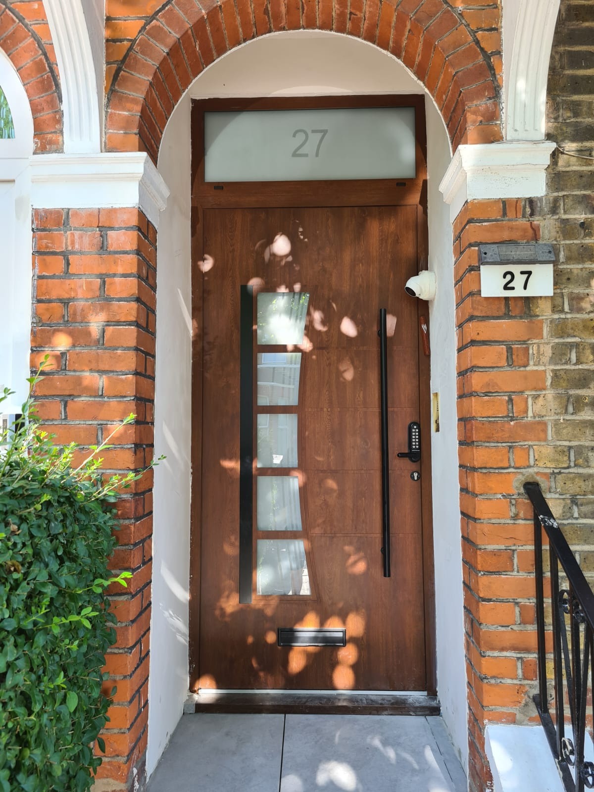 Nowoczesne, brązowe drzwi wejściowe z przeszkleniami i długim, czarnym uchwytem, numer domu '27' nad drzwiami i obok, kamera monitoringu widoczna nad drzwiami.